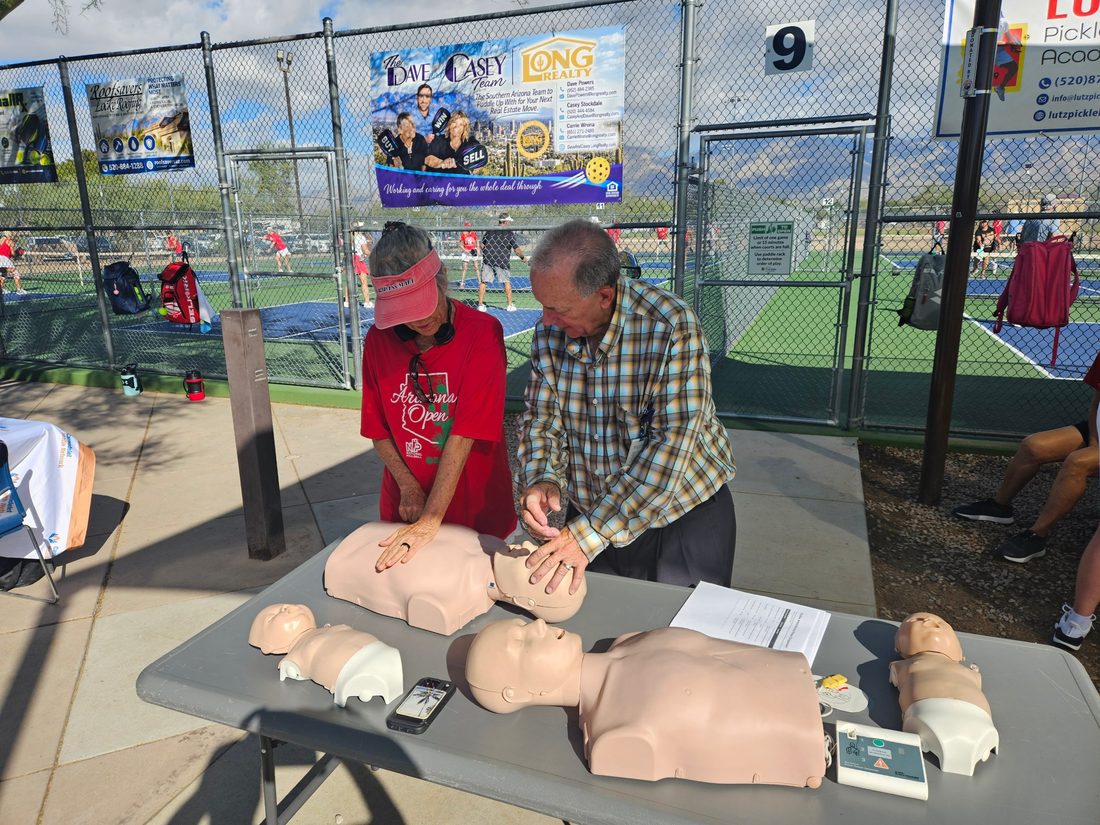 Two people practice chest compressions on CPR training manikins at a table beside the Udall Park pickleball courts.