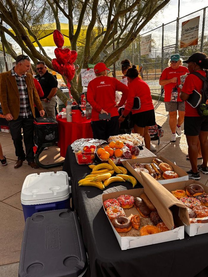 Players in red shirts gather under a shade canopy at Udall Park, with red heart balloons and a table of refreshments.
